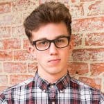 A young man with short brown hair and black glasses stands in front of a brick wall, wearing a red, white, and black plaid button-up shirt and holding a small pet costume, looking directly at the camera with a neutral expression.