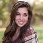 A young woman with long brown hair smiles at the camera, wearing a plaid shawl and holding a pet costume. The background is outdoors and blurred with green foliage.